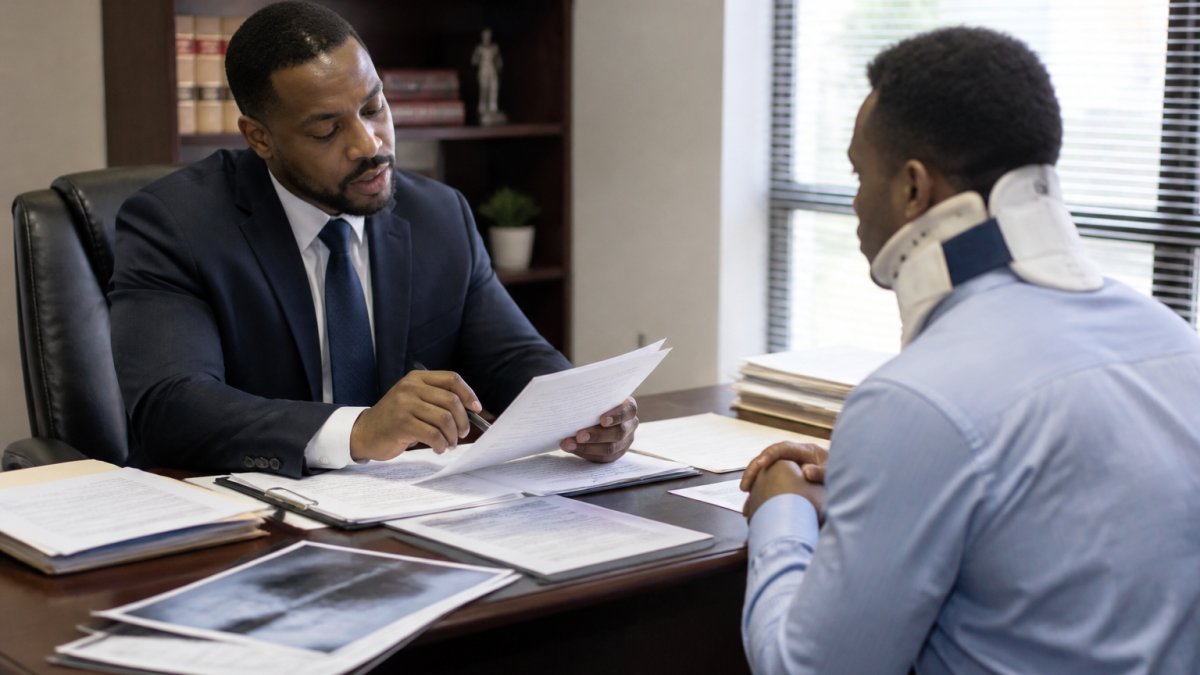 Professional meeting with car accident victim in office setting, reviewing settlement documents and medical records across desk with tablet and papers, natural lighting from window